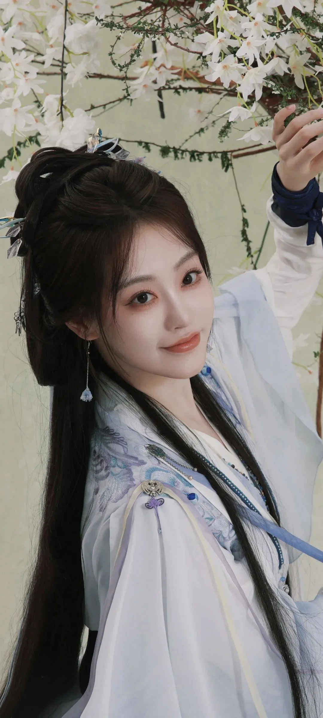 흰 꽃가지 아래에서 전통 의상을 입은 여성이 카메라를 향해 미소 짓고 있다.