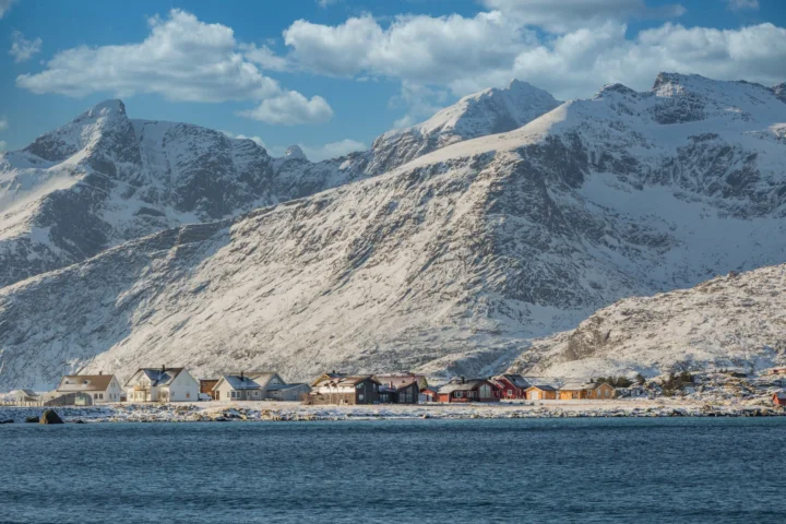白雪覆盖的群山矗立在海岸村落后方，前景是平静的蓝色海水和晴朗天空。