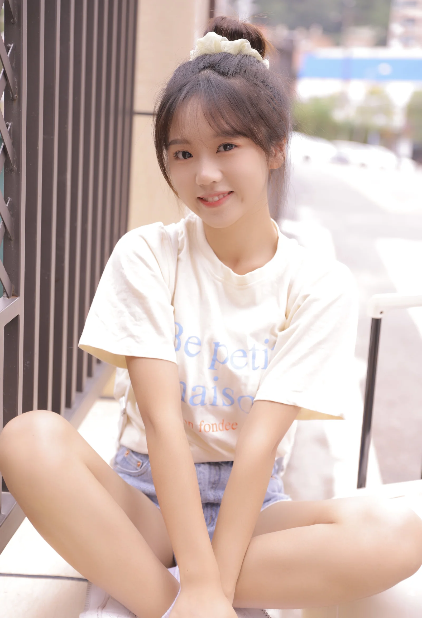 一位扎着发髻、身穿白色T恤的年轻女性坐在户外，对着镜头微笑。