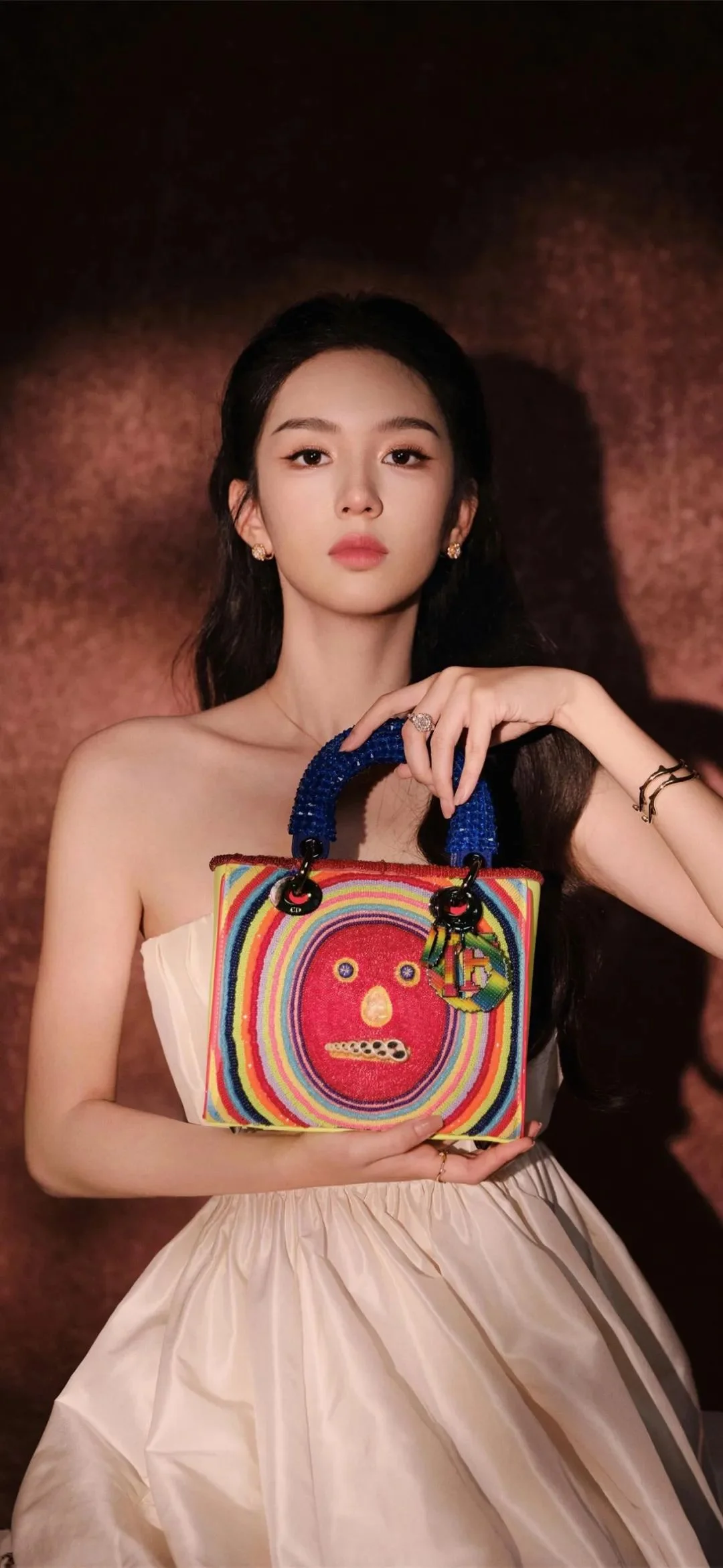 一位女性身穿浅色礼服，手持彩色手提包，在深色背景前摆姿势。