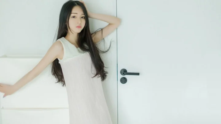 一名穿白色连衣裙的女子站在白色门旁摆姿势。