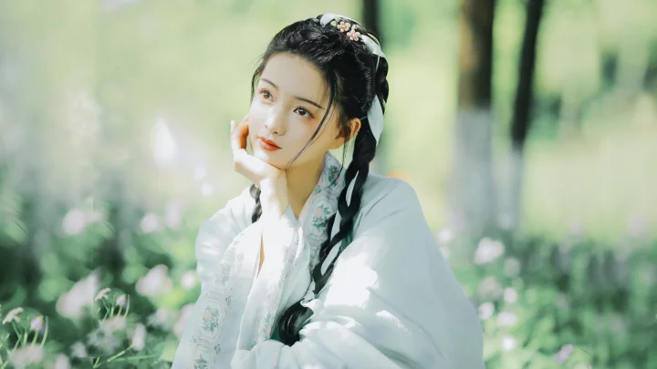 밝은 전통 의상을 입은 여성이 초록빛 숲속에서 옆모습으로 바라보고 있다.