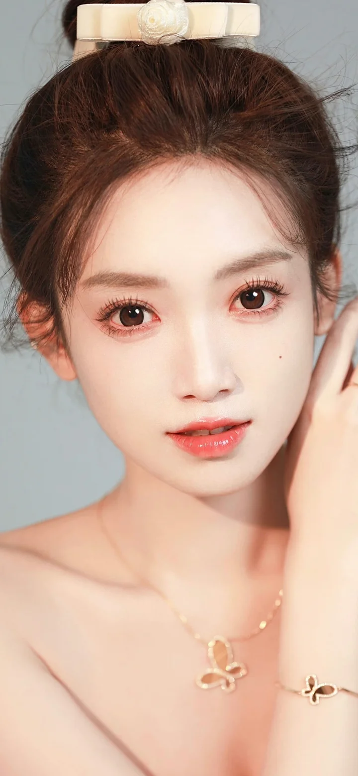 一名女性面对镜头展示盘发、精致妆容和项链，背景为浅灰色。