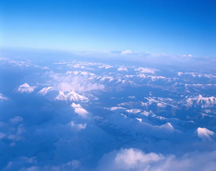从高空俯瞰绵延的雪山山脉，云雾与蓝色天空覆盖在远景之上。