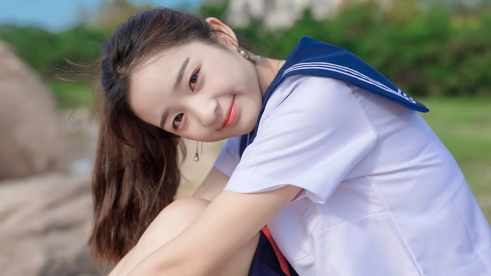 세일러풍 교복을 입은 여성이 야외에서 카메라 가까이에 앉아 미소 짓고 있다.