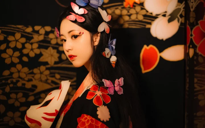 一名佩戴彩色蝴蝶发饰、身穿传统风格服饰的女子站在黑色花卉图案背景前。