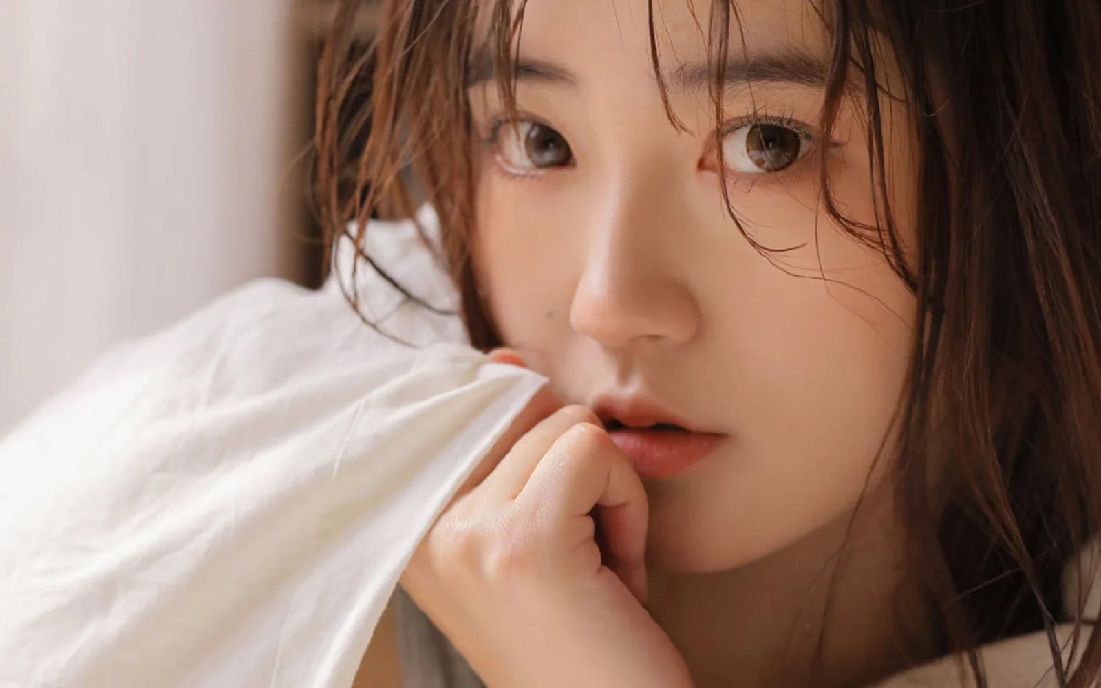 부드러운 빛 속에서 젖은 갈색 머리카락이 얼굴을 감싸고 손가락을 입술에 댄 여성의 클로즈업 인물 사진이다.