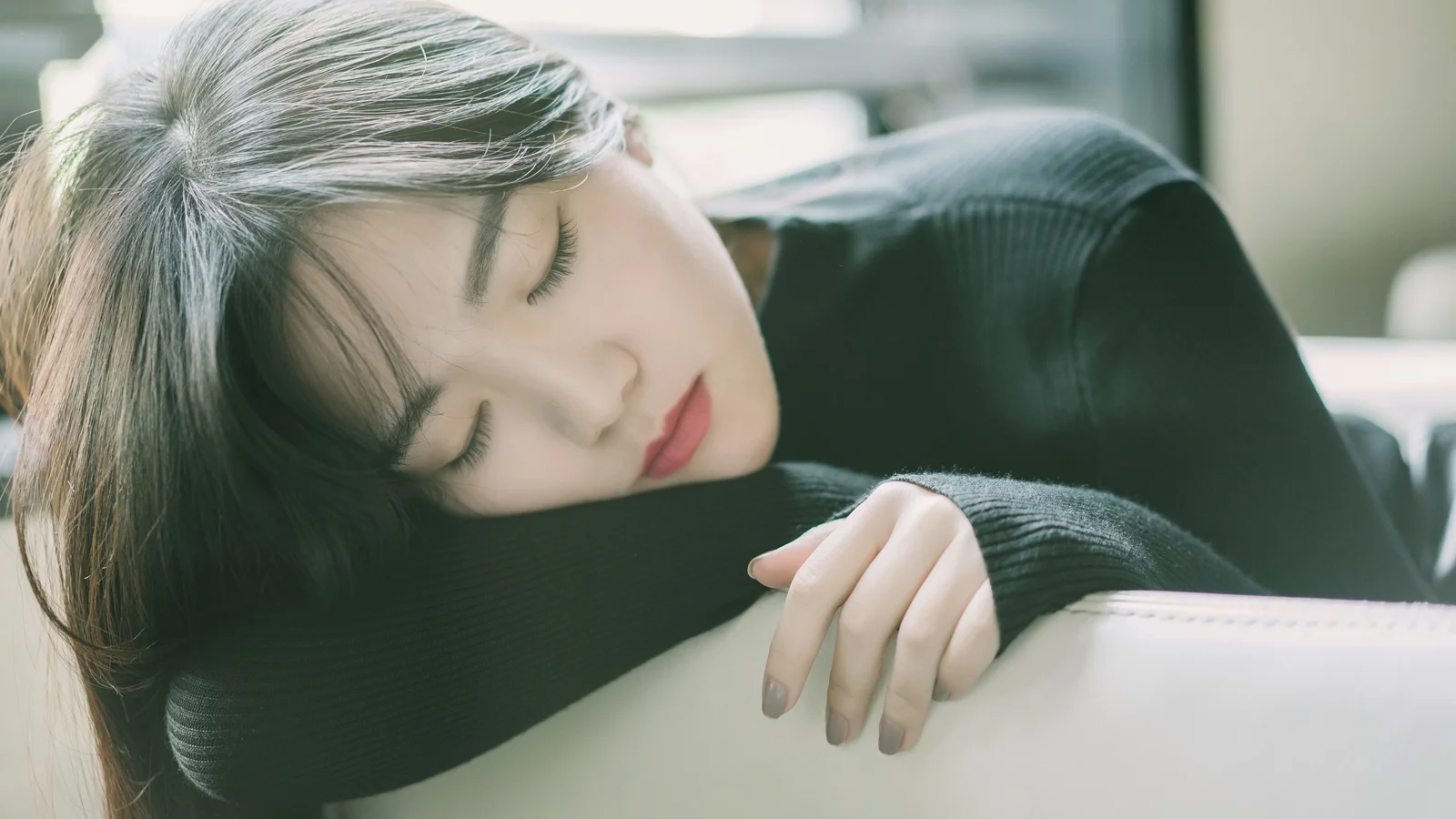 一名黑髮女子穿著黑色毛衣，閉眼靠在白色表面上休息。