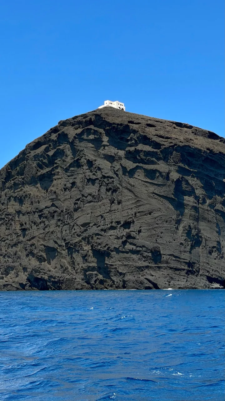 蓝色海水映衬着陡峭的深色火山岩壁，山顶可见一座白色建筑，天空晴朗无云。