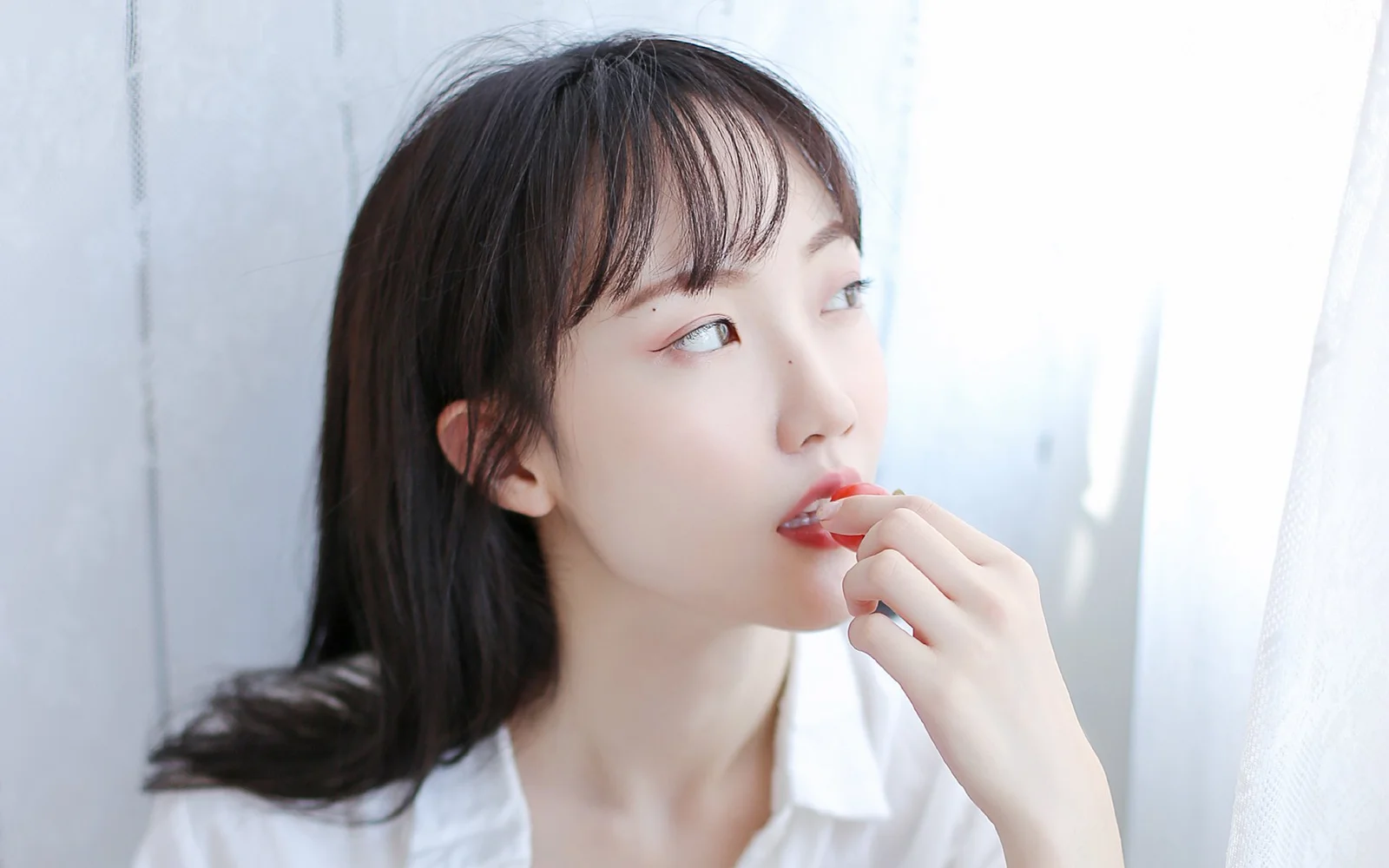 一名女性站在窗邊，手持唇彩靠近嘴唇，柔和的自然光照亮她的側臉。