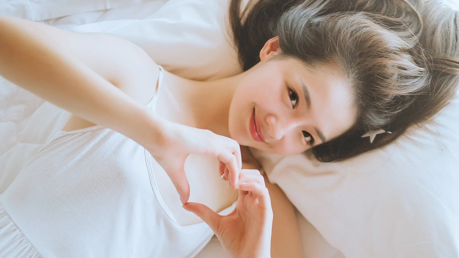 一名成年女性躺在白色床上，用双手在胸前摆出爱心手势。