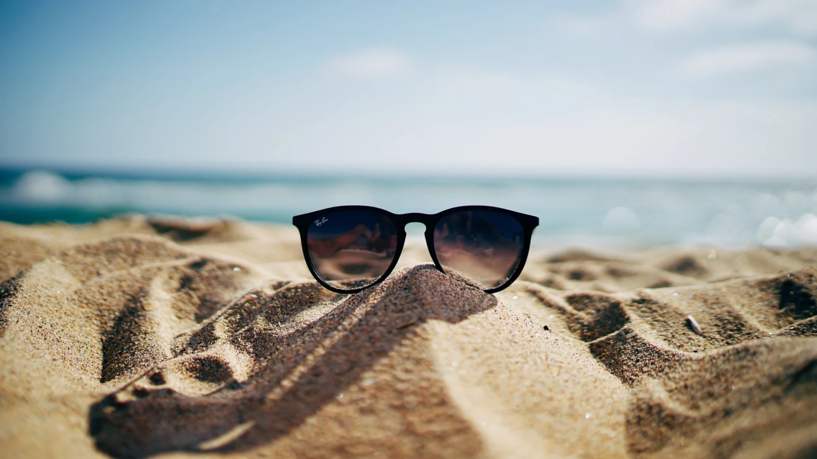 Sunglasses resting on a sandy beach with a softly blurred ocean and shoreline in the background.