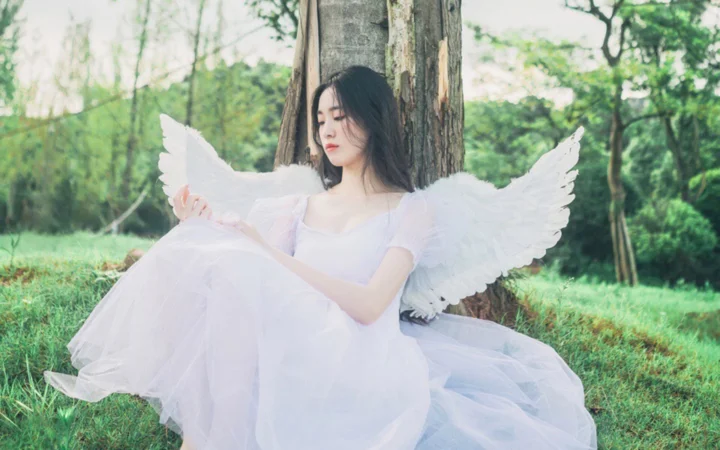 흰 드레스와 흰 깃털 날개를 착용한 여성이 숲속 나무 옆에 앉아 있다.