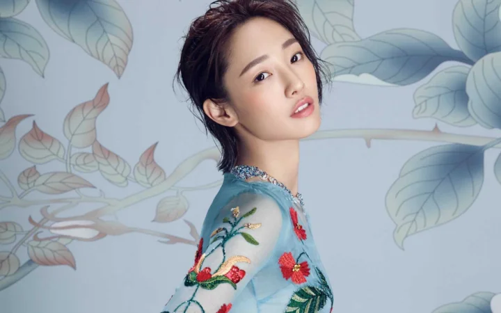 一位成年女性身穿蓝色刺绣服饰，站在花叶图案背景前面向镜头。