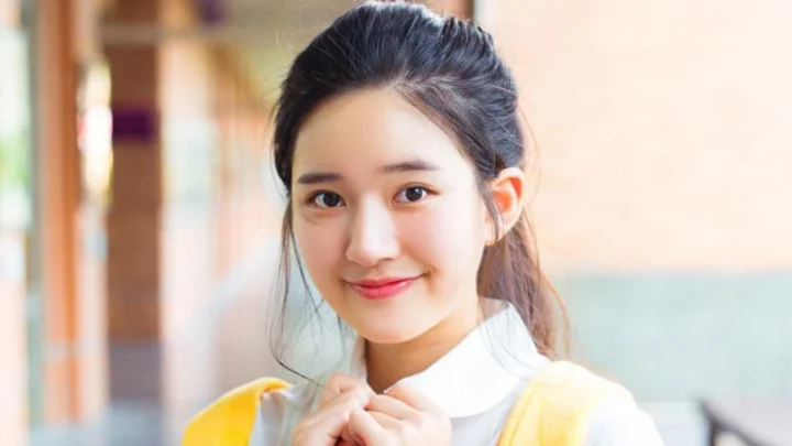 一位面带微笑的女性身穿白色领口上衣和黄色衣袖，在室内背景前望向镜头。