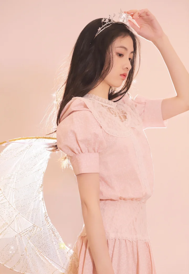ピンクのドレスときらめく髪飾りを身につけ、翼のような装飾を背負った女性がピンクの背景の前に立っています。