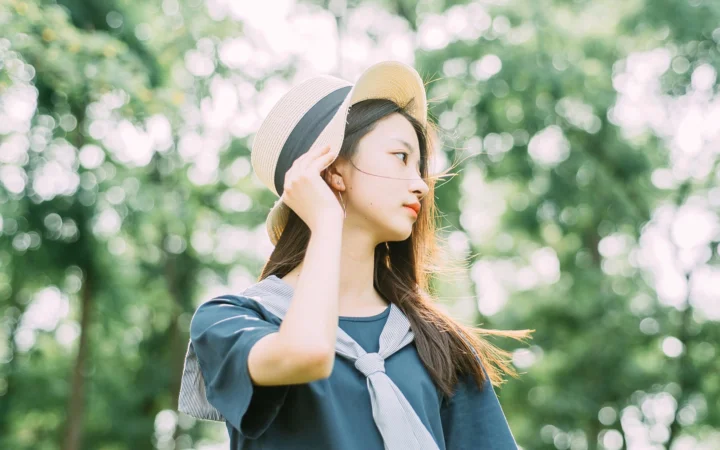 明るい色の帽子をかぶった女性が、木漏れ日の緑を背景に立っている。