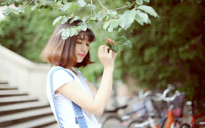 一名年轻女子站在户外树枝下，手持一片叶子，背景中可见自行车。