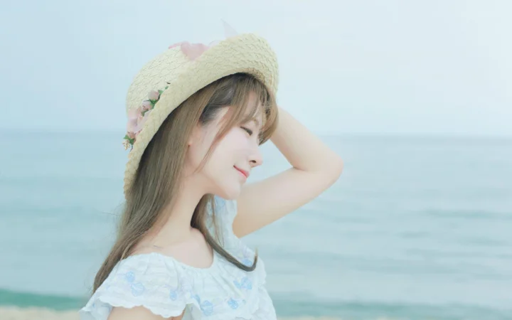 麦わら帽子をかぶった女性が、穏やかな青い海を背景に海辺に立っている。