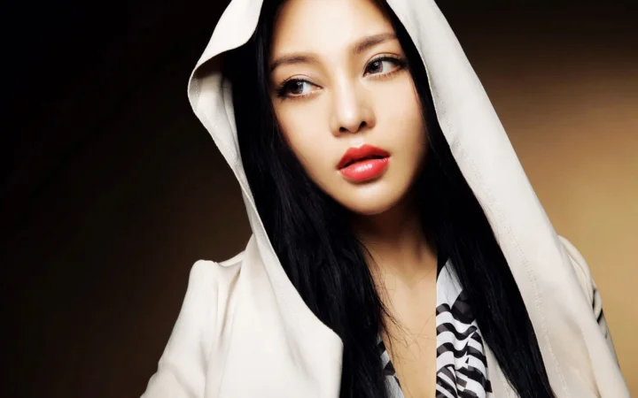 흰색 후드 의상을 입은 긴 머리 여성이 어두운 배경 앞에서 옆을 바라보고 있습니다.