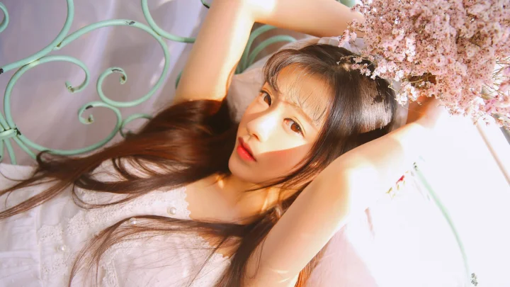 一名长发女子躺在柔和的阳光下，身旁映着粉色花朵与浅绿色装饰。