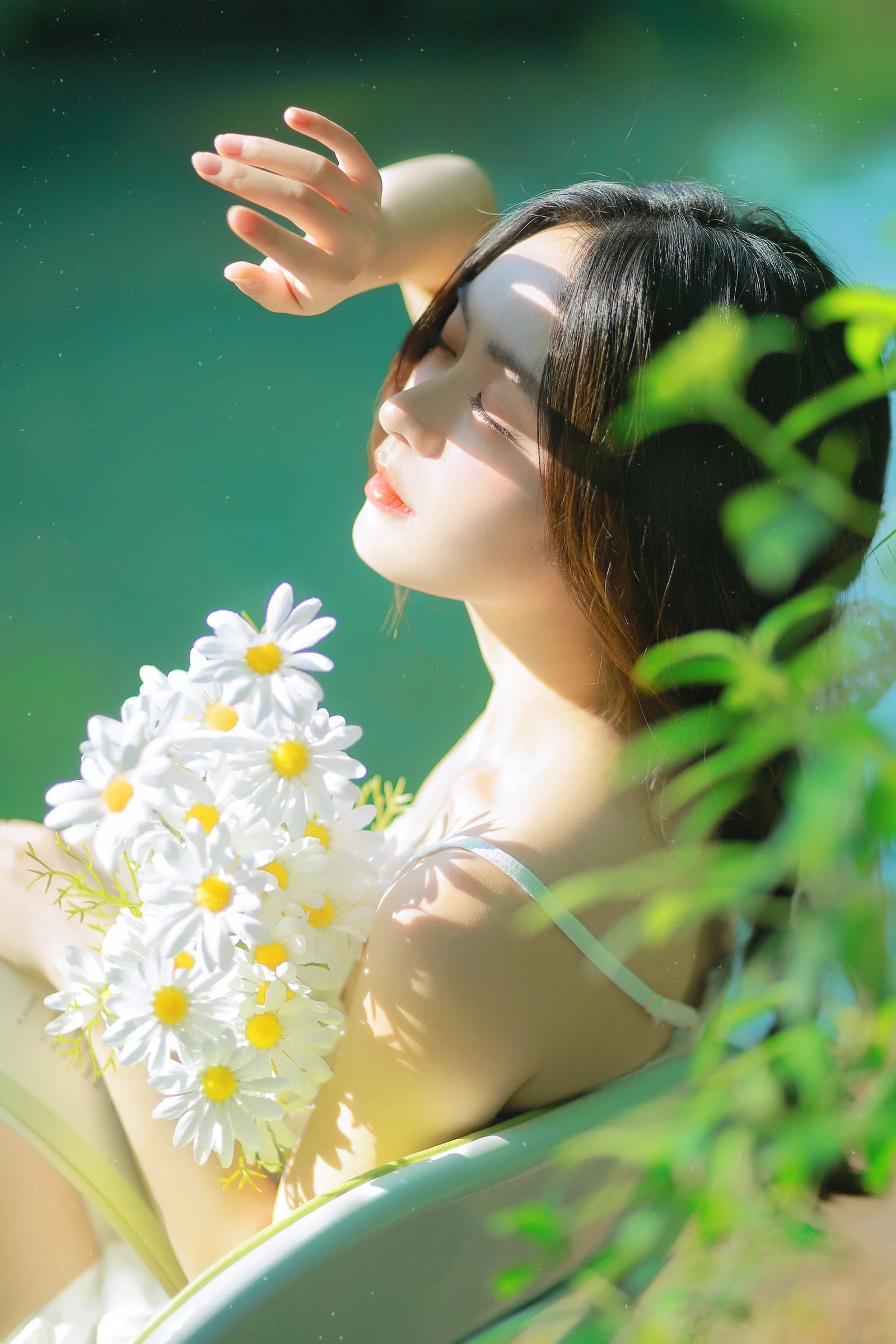 한 여성이 햇살과 푸른 잎사귀에 둘러싸여 흰 데이지 꽃다발을 안고 있다.