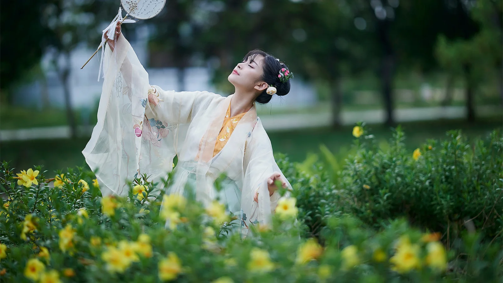 一名身穿传统服饰的女子站在户外花丛中，手持装饰物并仰头望向上方。