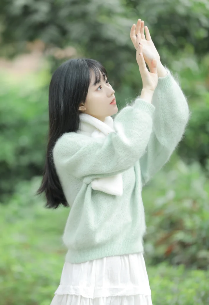 一名长发女子穿着白色毛衣站在林间，双手向上举起。