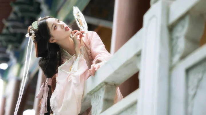 一名身穿粉色传统服饰的女子倚在白色栏杆旁，手持团扇望向远处。