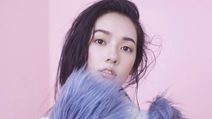 一名成年女性在粉紫色背景前被柔和光线照亮，前景环绕着紫色毛绒材质。