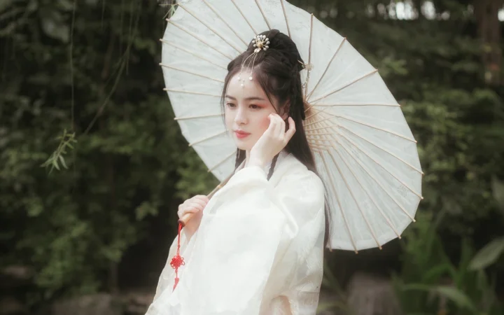 흰 옷을 입은 여성이 야외에서 흰 종이우산을 들고 흐릿한 초록 숲을 배경으로 서 있다.