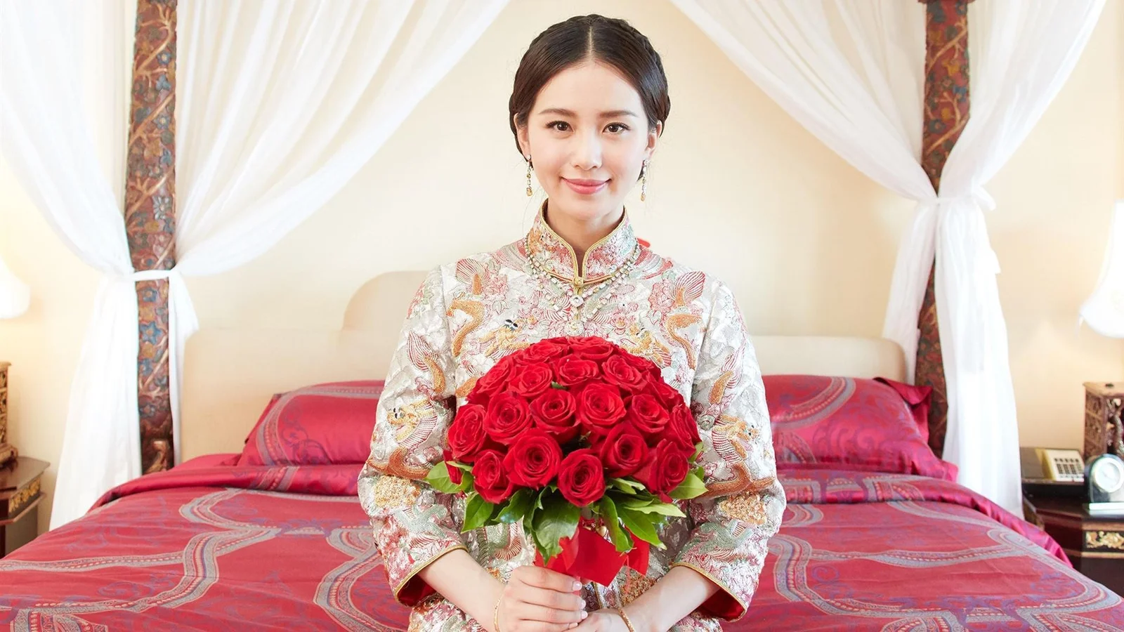 전통 의상을 입은 여성이 침대 앞에 앉아 빨간 장미 꽃다발을 들고 있다.
