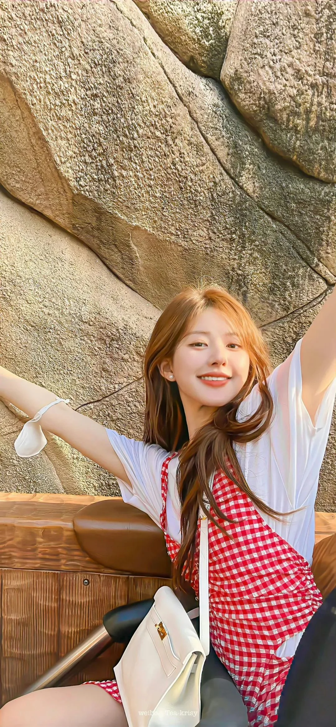 긴 머리의 여성이 바위 배경 앞에 앉아 빨간색과 흰색 체크무늬 원피스와 흰색 상의를 입고 있다.
