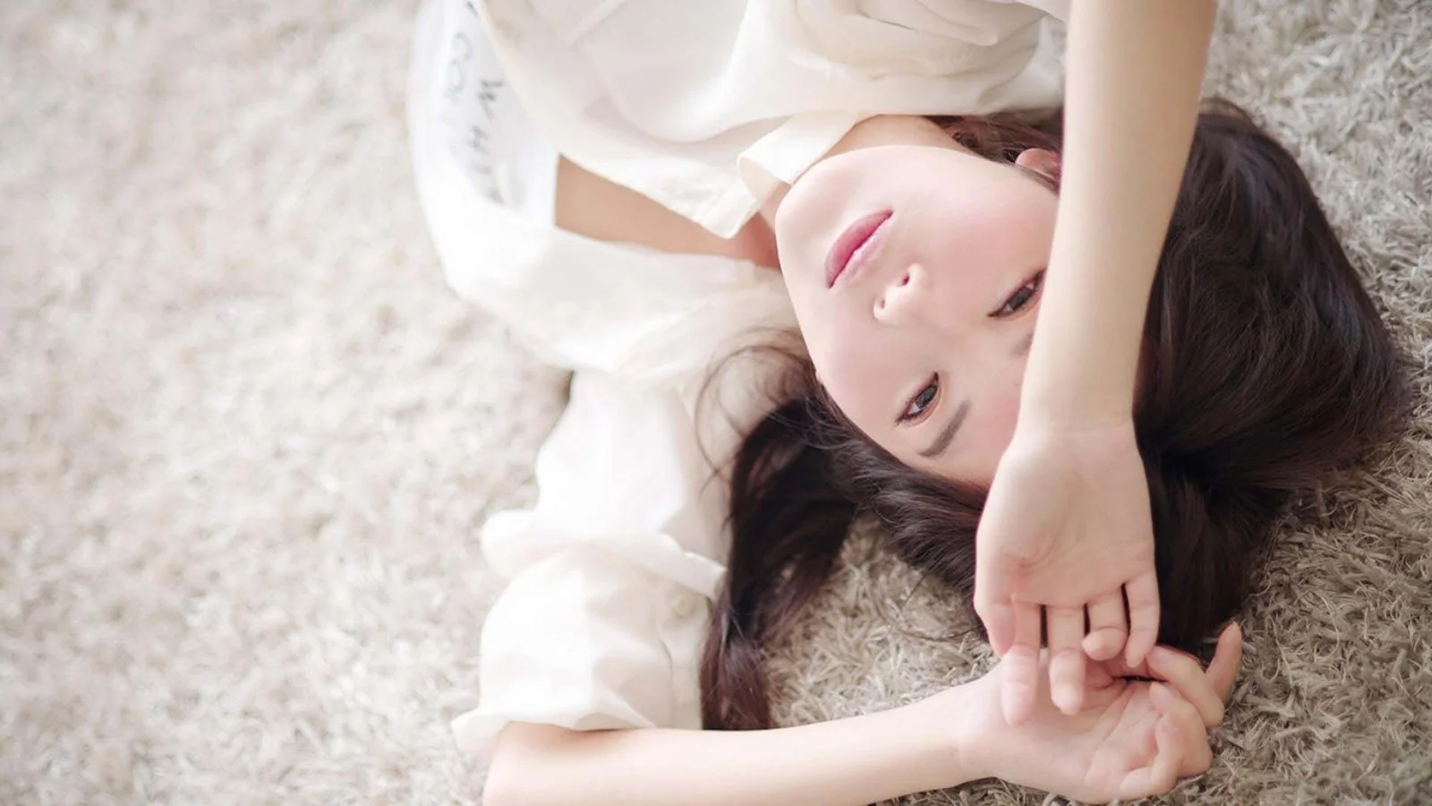 一名身穿白色衣服的女子躺在柔软地毯上，手臂举过头顶看向镜头。