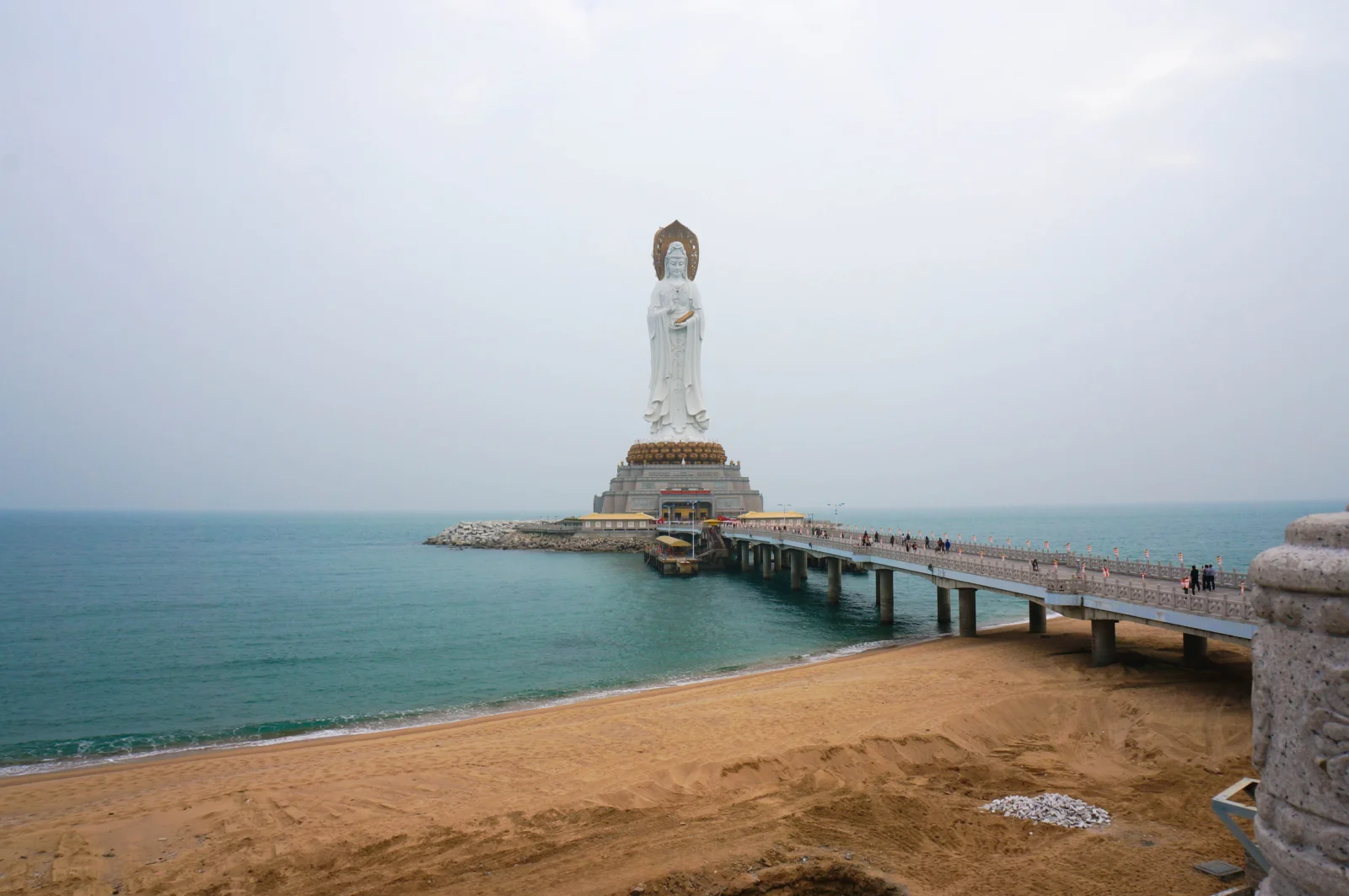 沙灘、海水與棧橋盡頭的白色雕像在朦朧天空下構成寧靜的海岸景色。