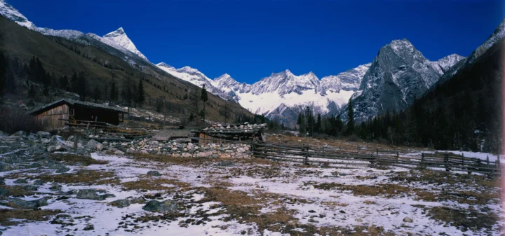 积雪覆盖的高山山谷在深蓝天空下延展开来，前景可见木屋和围栏。