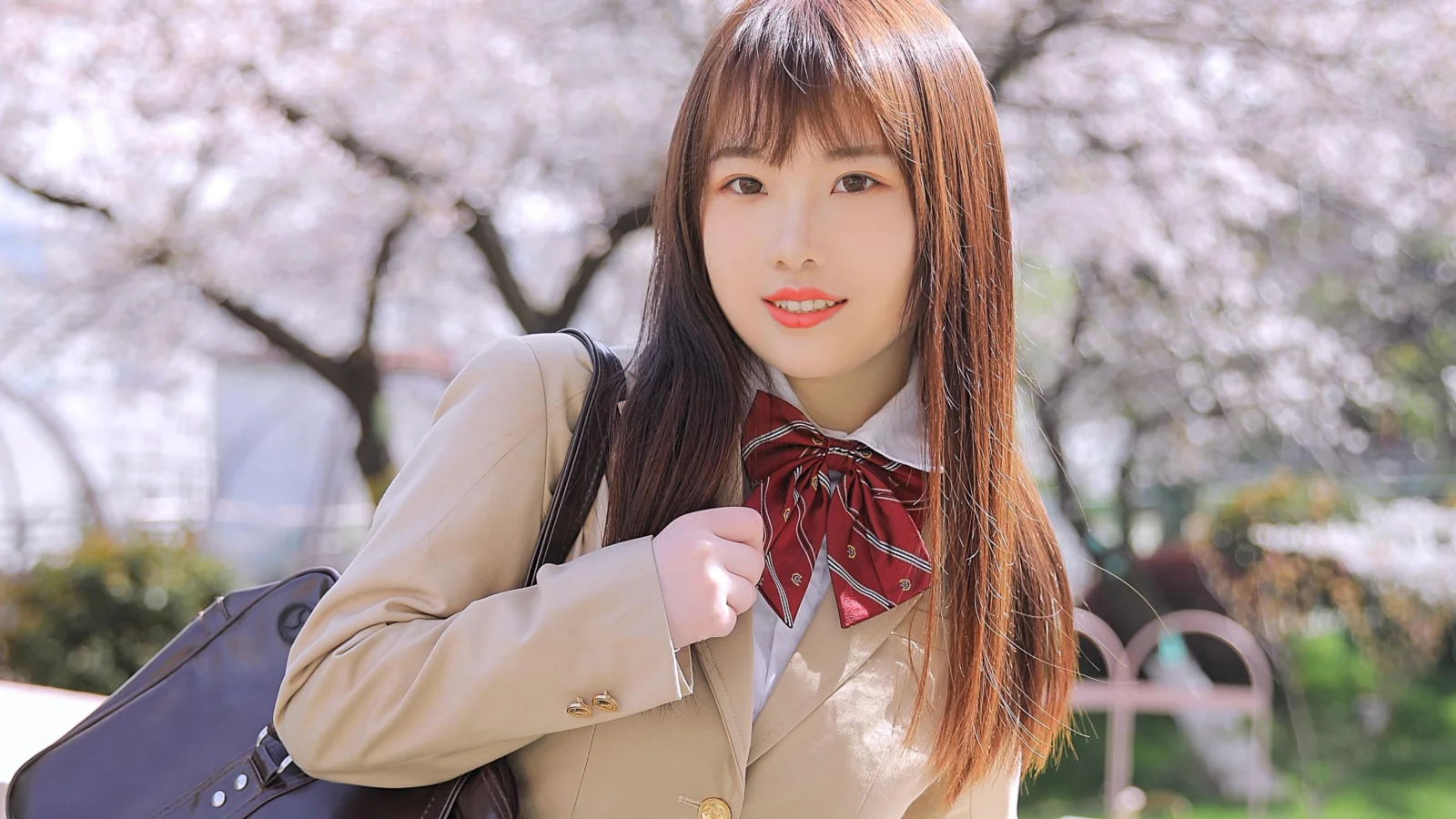 긴 머리의 여성이 밝은색 재킷을 입고 가방을 멘 채 만개한 벚꽃나무 아래 서 있다.