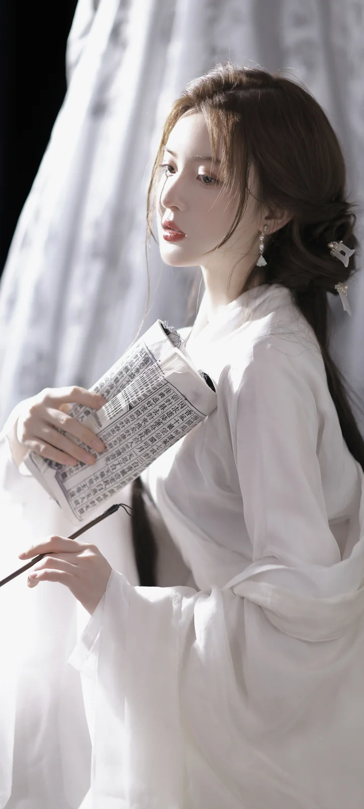 白い長袖の服を着た女性が窓辺で新聞を手にしている。
