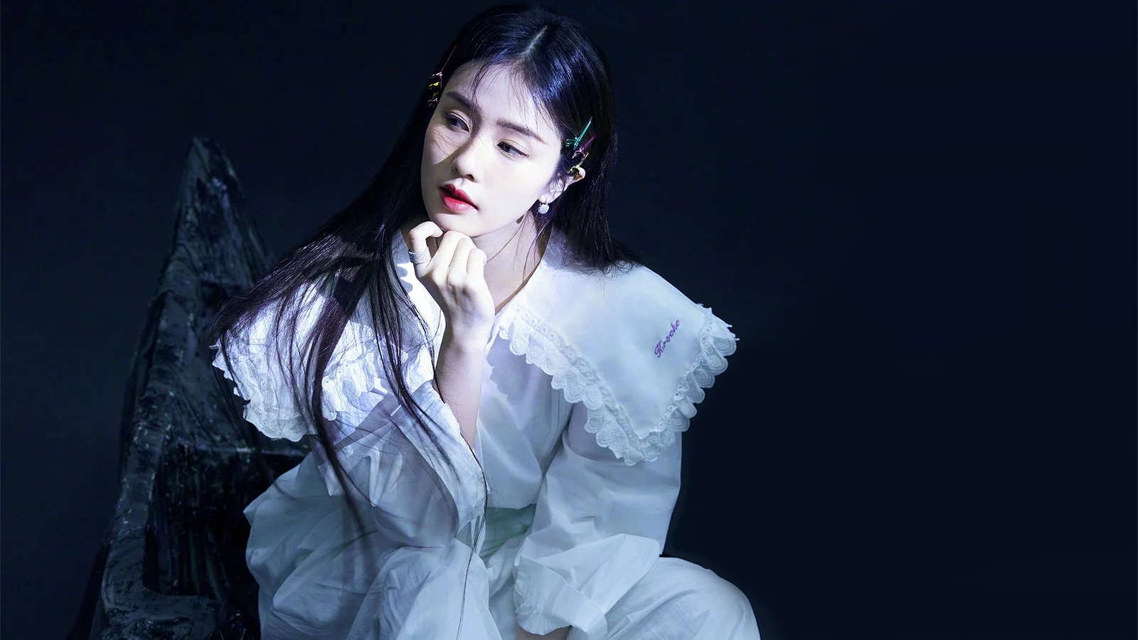 긴 머리의 여성이 흰 드레스를 입고 어두운 배경 앞에서 옆을 바라보고 있다.