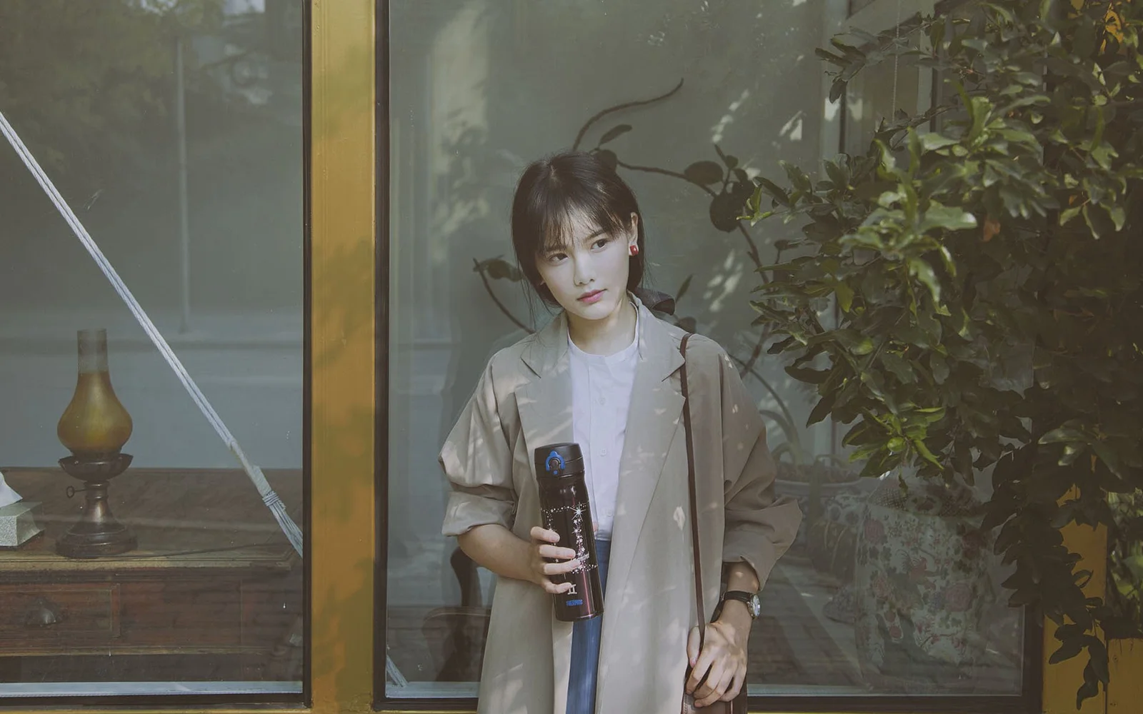 한 여성이 창가에 서서 물건을 들고 있으며 옆에는 초록 식물이 보입니다.