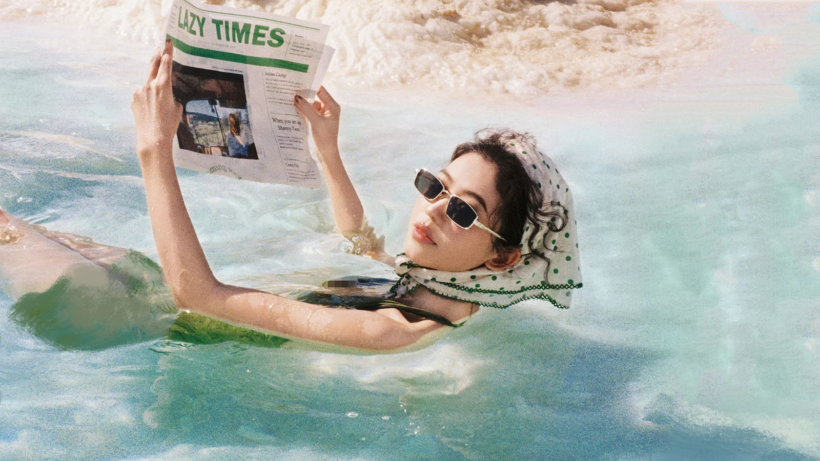 サングラスをかけた成人女性が、水に浮かびながら新聞を手にして読んでいる。