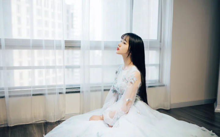 一名身穿白色连衣裙的女子坐在明亮窗边，透过纱帘沐浴在柔和的室内光线中。