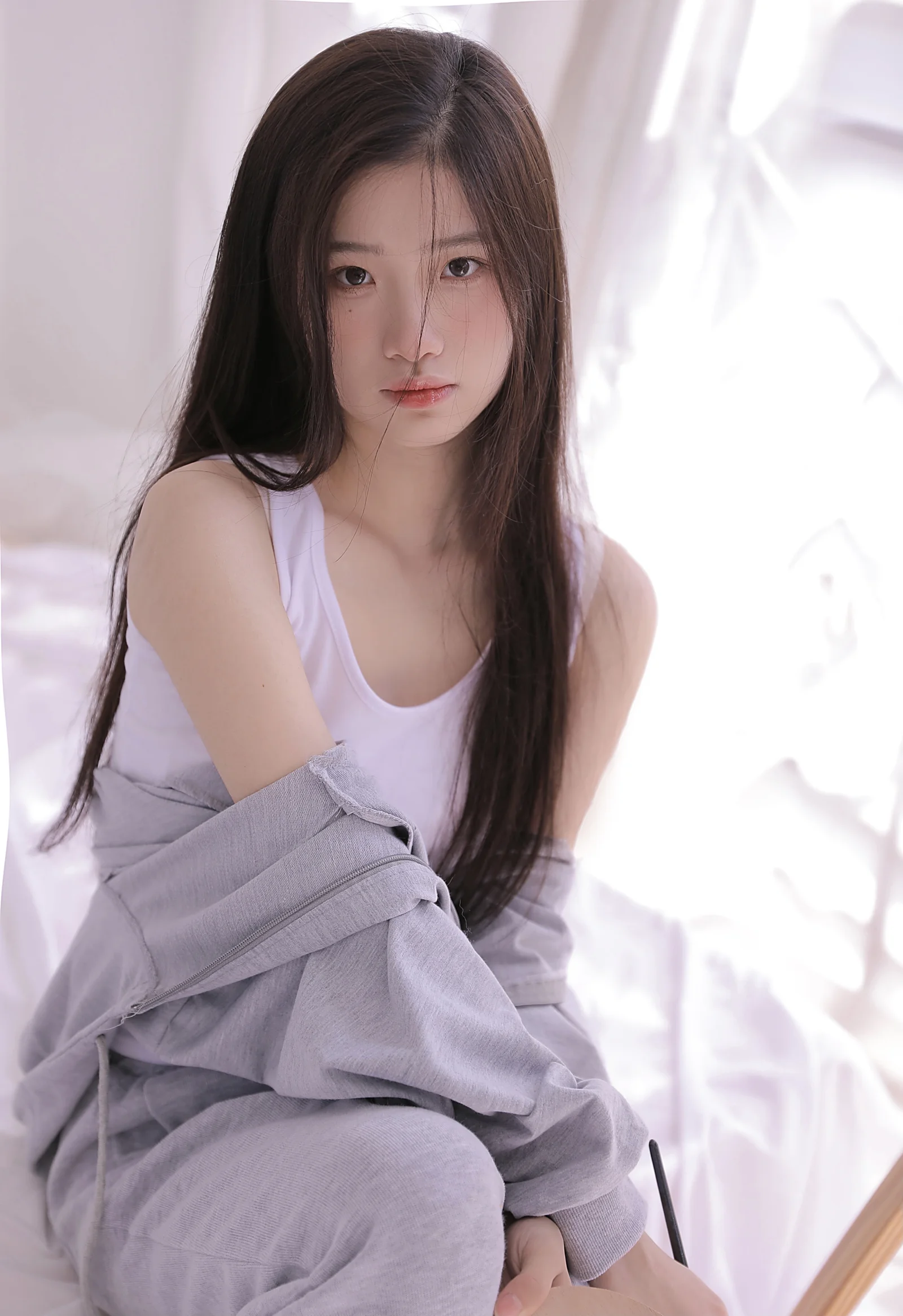 明るい窓辺で淡い色のカジュアル服を着た長髪の女性がカメラを見つめるポートレート写真です。