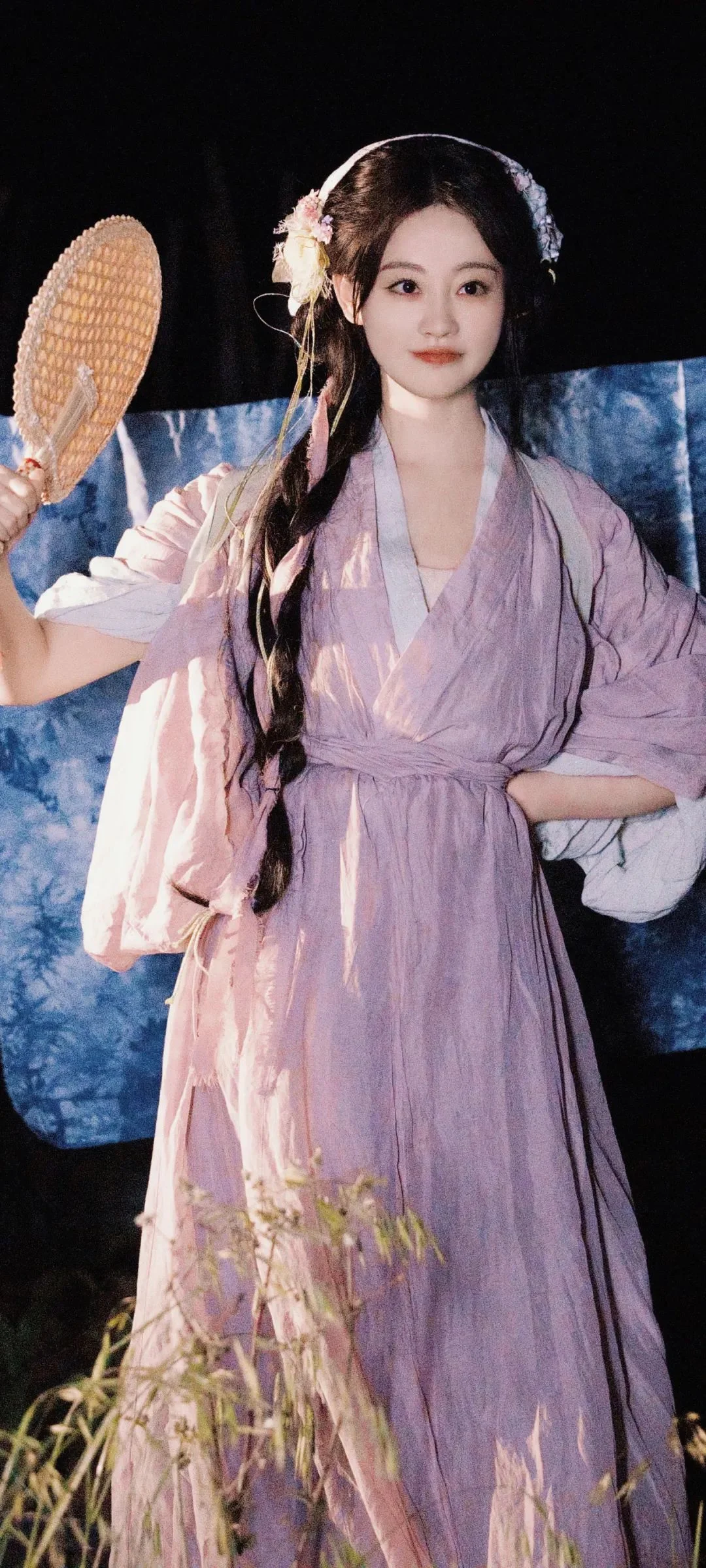 一名身穿淡紫色传统服饰、手持团扇的女子站在深色背景前。