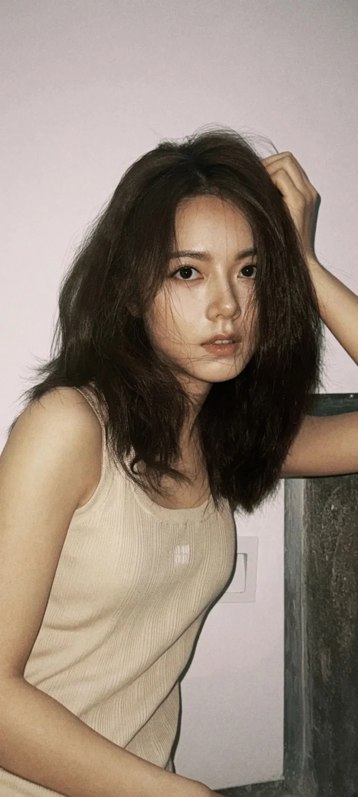 一名长发女性身穿浅色背心，在室内靠墙摆姿势。