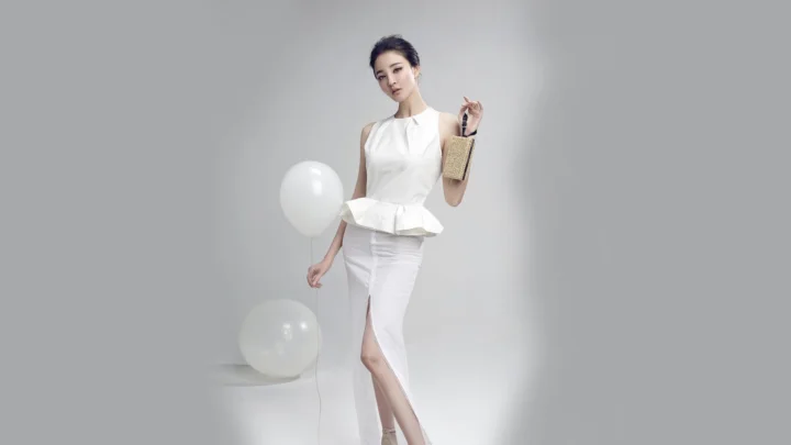 一位身穿白色服装、手提编织包的女性站在灰色背景前，身旁摆放着白色气球。