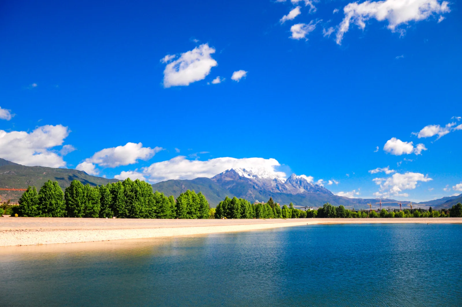 蓝天下的湖泊、绿树与远处雪山构成宁静的自然风景。