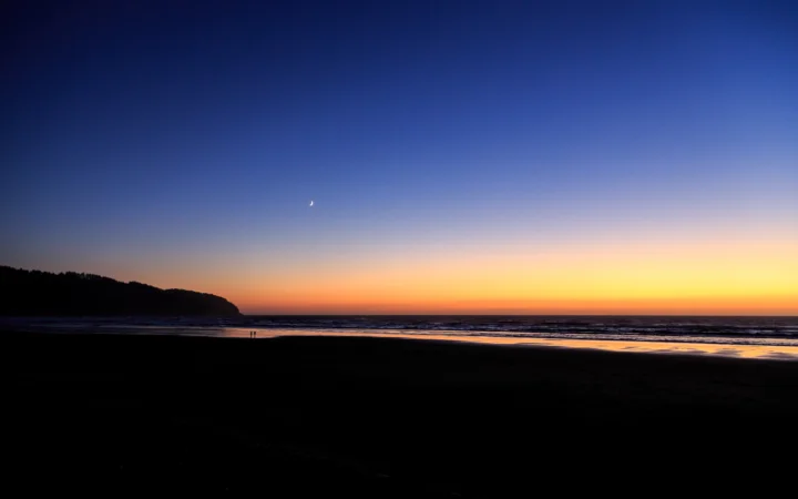 暮色中的海滩延伸至海面，远处低矮海岬映衬着橙黄色地平线与深蓝天空，天空中可见一弯月牙。