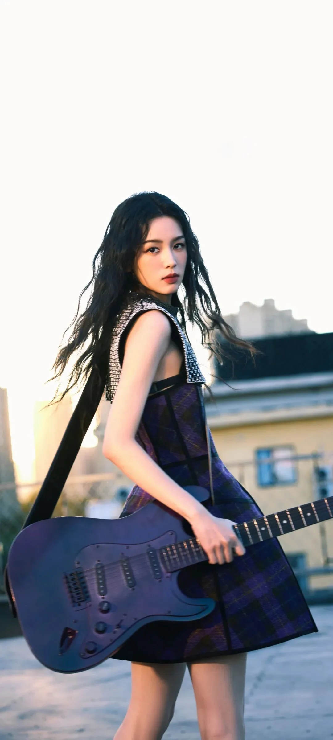 一名女子在户外手持电吉他面对镜头，背景可见建筑物。