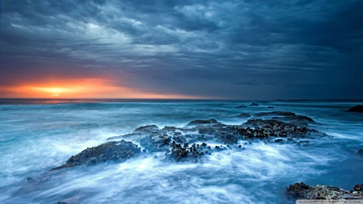 夕阳映照着海平面，海浪在前景的礁石间翻涌。